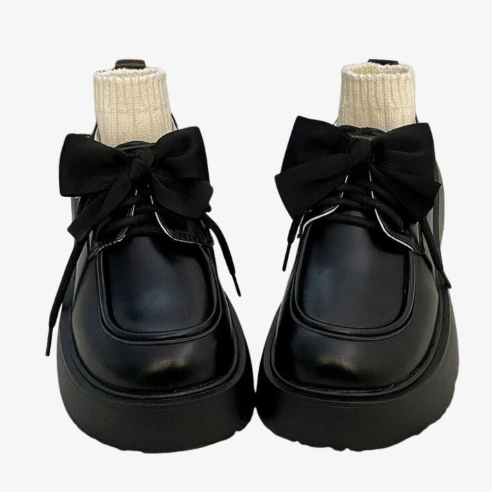Ein Paar Mokassins aus schwarzem Glattleder auf einem weißen Bett, eine große Schleife schmückt das Oberteil und kleine beigefarbene Socken füllen den Schuh.