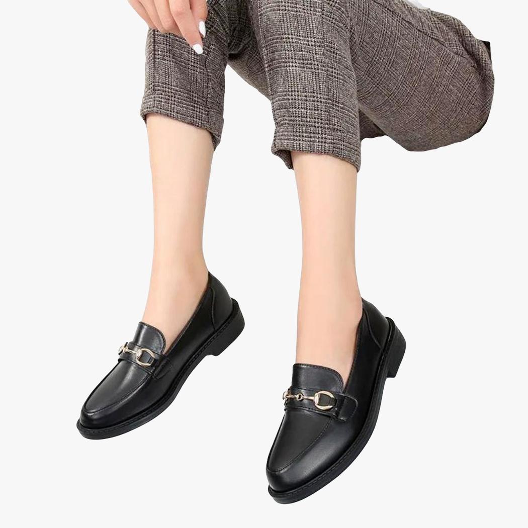Zu sehen sind die Füße einer sitzenden Frau, die schwarze Loafer mit Metallschnalle und Blockabsatz trägt