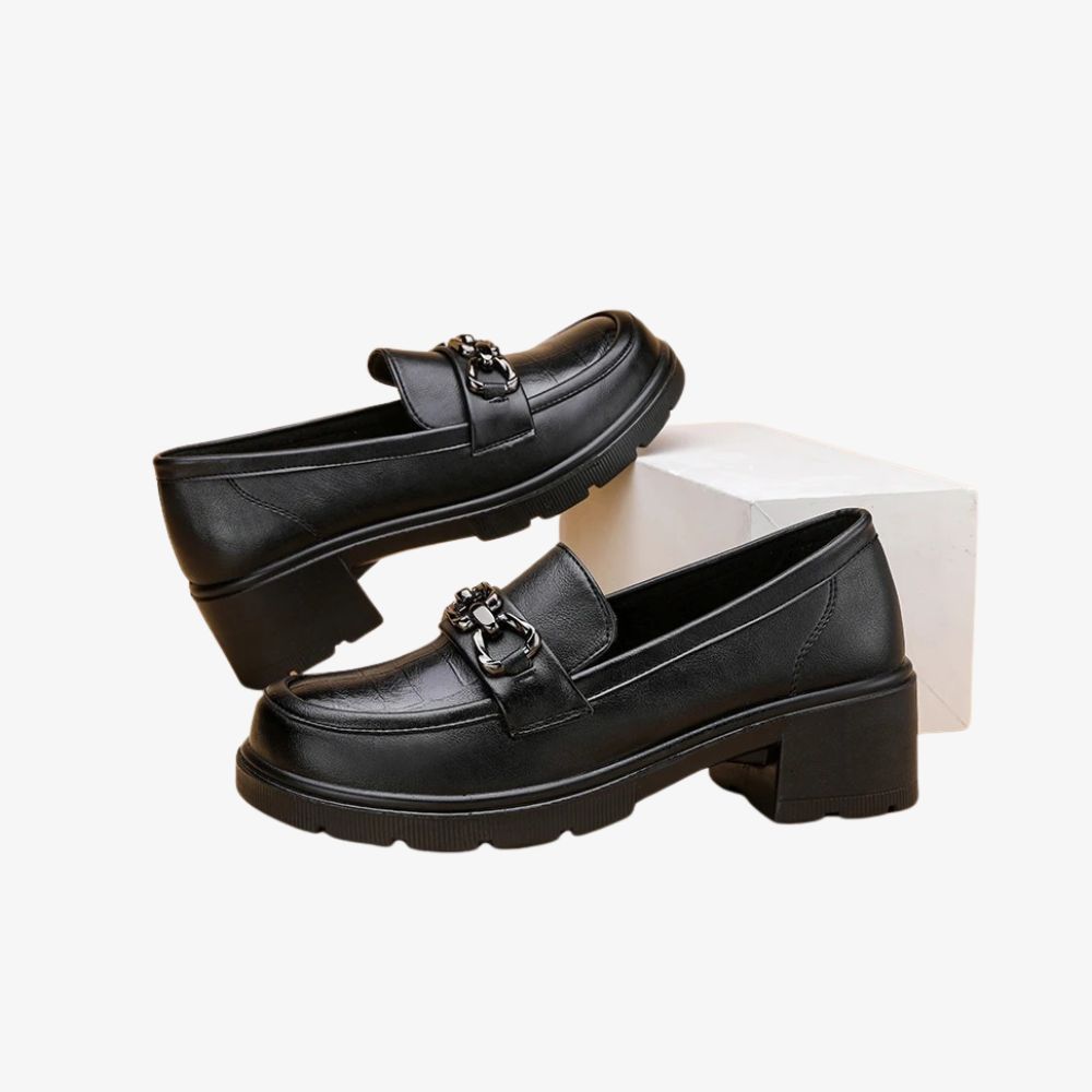 Ein Paar schwarze Mokassins mit Absatz, ein Schuh steht auf einem weißen Podest.