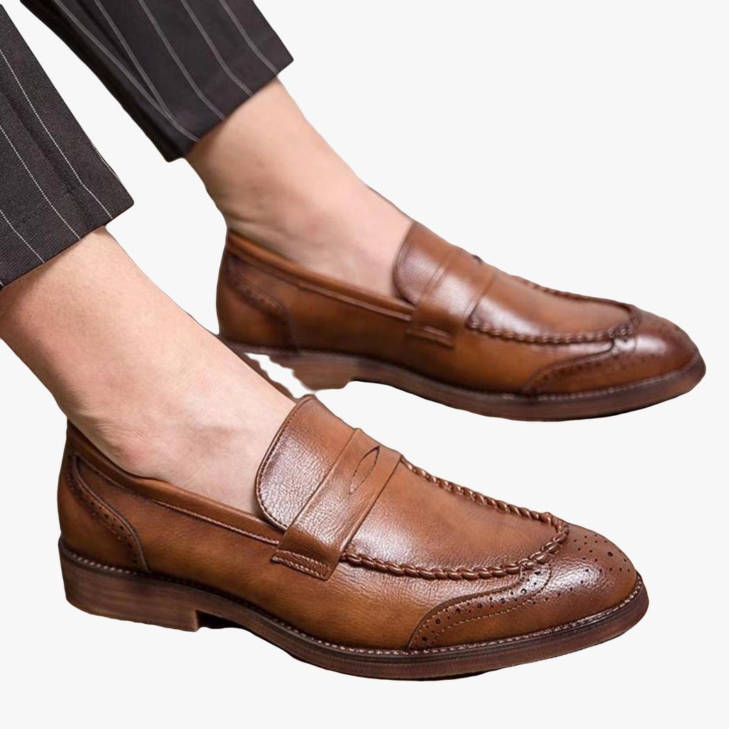 Zu sehen ist ein sitzender Mann, man sieht seine Füße, und er trägt braune Loafer aus Leder