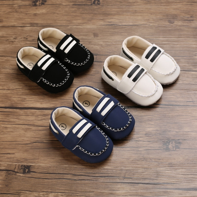 Auf braunem Parkett, drei Paar Baby-Boot-Mokassins mit Steppnähten und zwei weißen Balken auf der oberen Zunge. Sie sehen ein weißes, blaues und schwarzes Paar