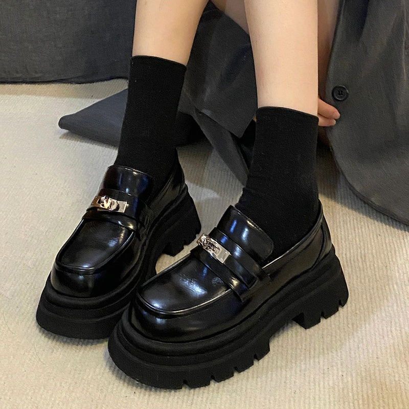 Schwarze Mokassins aus glänzendem Glattleder mit einer dicken Sohle an den Füßen einer Frau, die auf dem Boden sitzt und kurze schwarze Socken trägt.