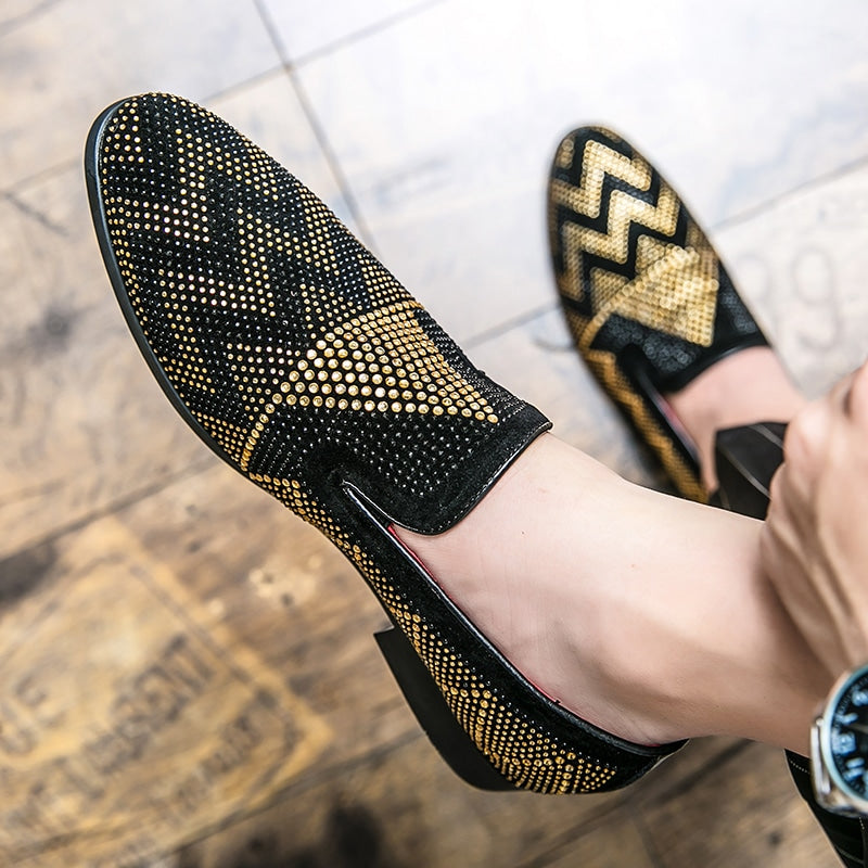 Barfuß getragenes Paar Mokassins in Schwarz mit geometrischen Stickereien aus goldenen Strasssteinen. Ein Fuß steht auf dem Boden auf einem braunen Parkettboden.