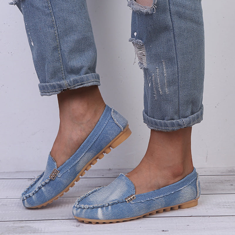 Ein Paar weiche Mokassins aus verwaschenem blauem Denim mit beigefarbener Gummisohle mit Noppen, barfuß getragen von einer Frau in zerrissenen blauen Jeans mit sichtbaren Knöcheln