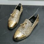 Auf einem grau marmorierten Boden, ein Paar Mokassins mit Quasten aus Lack und goldenen Pailletten mit schwarzen Schuhspannern an der Innenseite