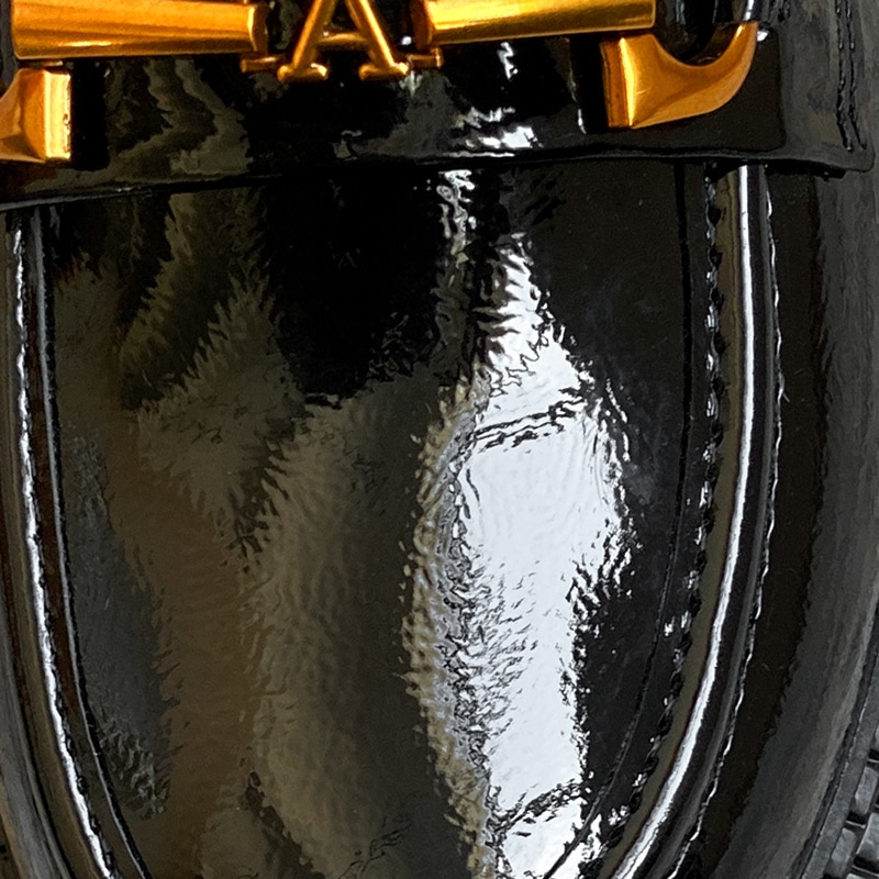 Foto eines Zooms auf einen schwarzen Mokassin aus Lackleder mit einer goldenen Kette und einem A