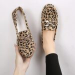 Ein Paar Mokassins im Leopardenmuster mit Ziernähten und silbernen Strassringen auf der Oberseite. Ein Schuh steht in einem Fuß und der andere wird in einer Hand gehalten.