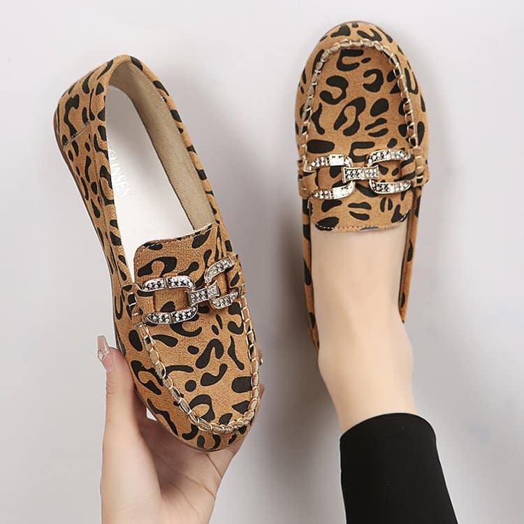 Ein Paar abgerundete Mokassins mit braunem Leopardenmuster und silbernen Strassringen, ein Schuh am Fuß mit einer schwarzen Strumpfhose und der andere in einer Hand gehalten