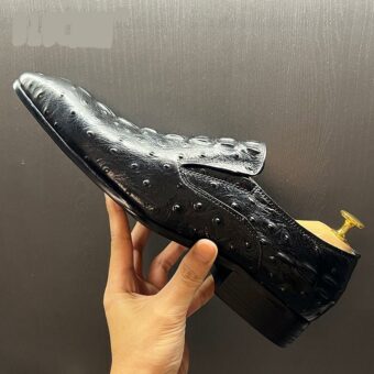 Eine Person hält einen schwarzen krokodilartigen Mokassin mit einem Schuhanzieher in der Hand