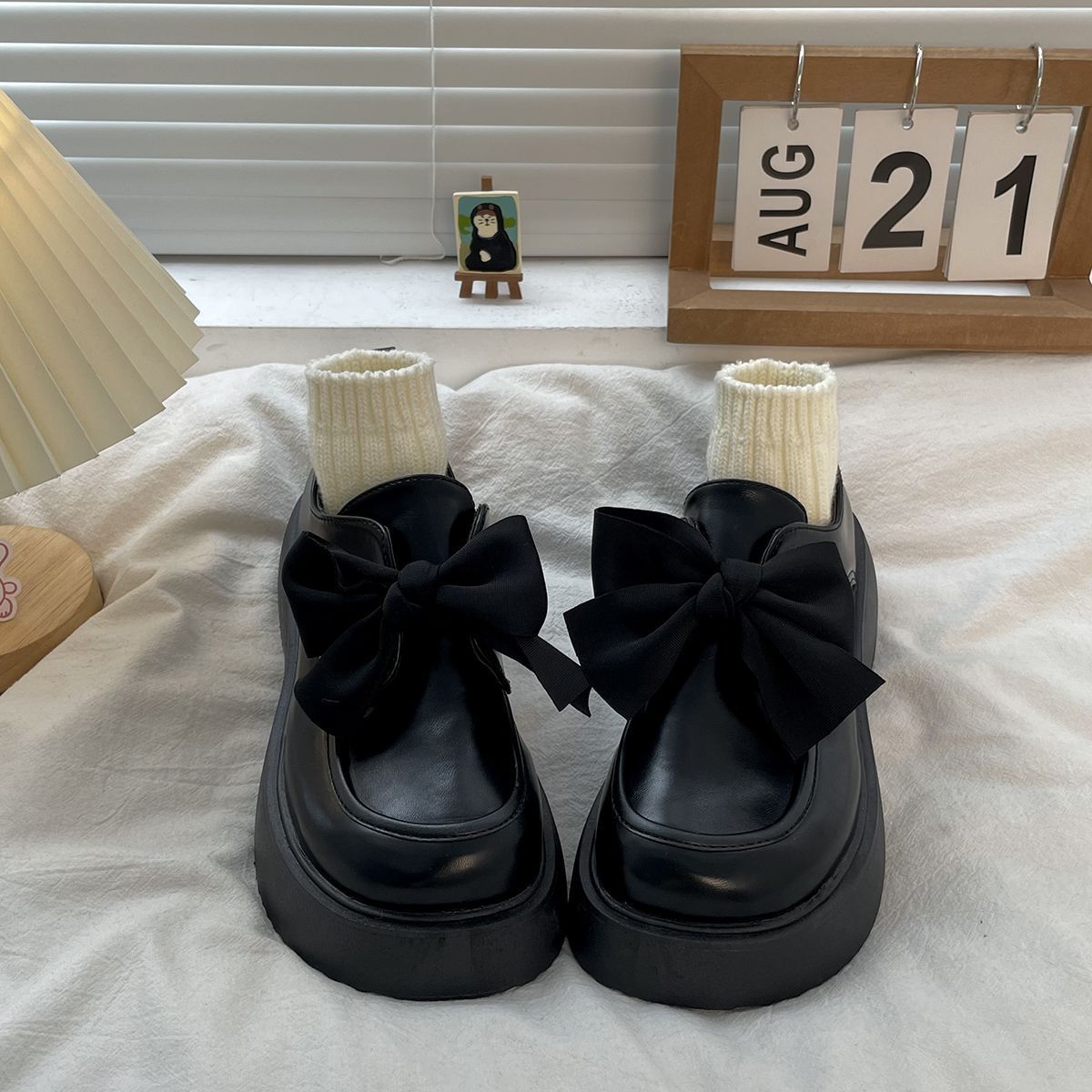 Ein Paar Mokassins aus schwarzem Glattleder auf einem weißen Bett, eine große Schleife schmückt das Oberteil und kleine beigefarbene Socken füllen den Schuh.