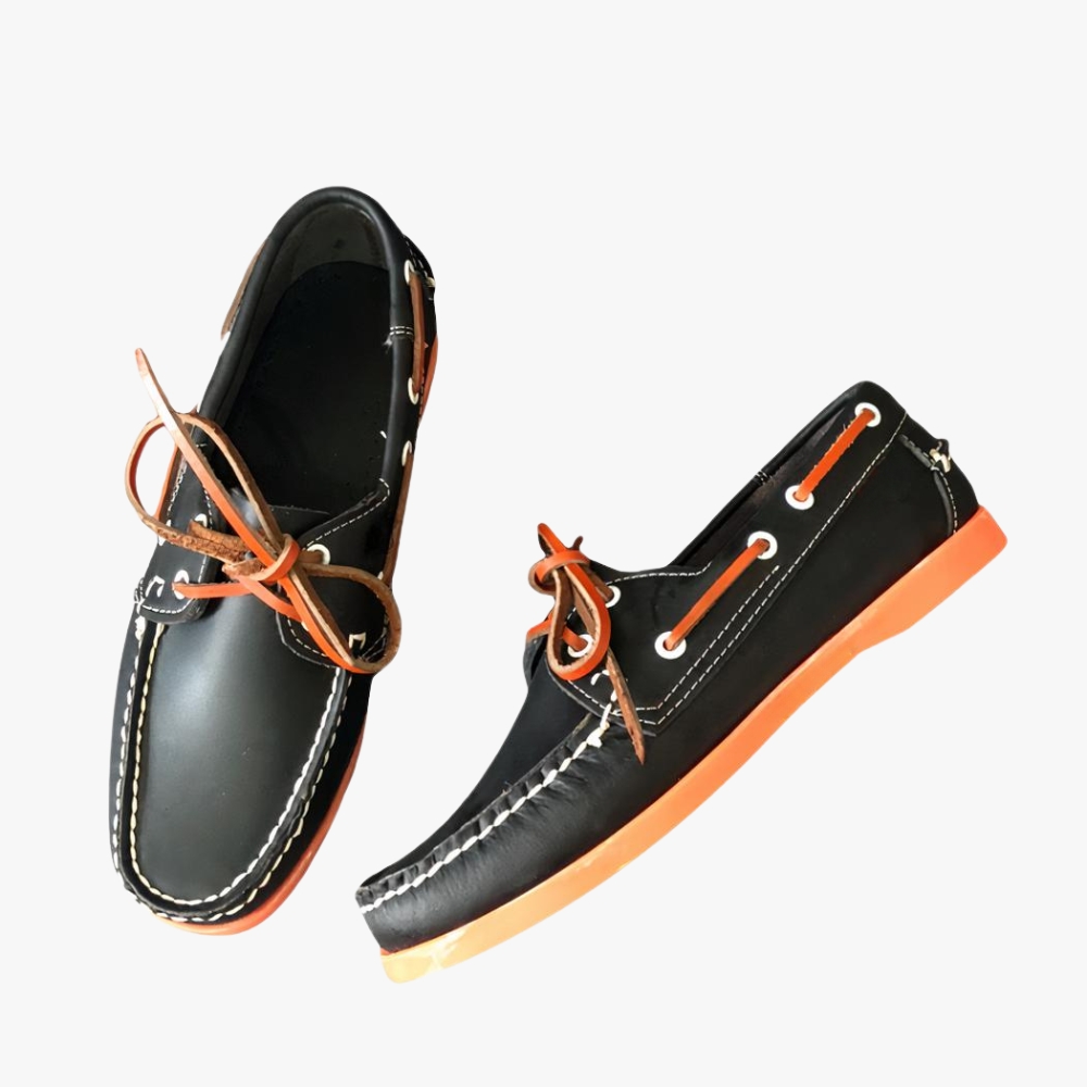 Schwarze Bootsmokassins mit orangefarbener Sohle, auf einem grauen Sofa liegend