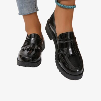 Paire de mocassins noirs vernis avec franges portés par une femme avec un jean bleu et un bracelet à la cheville.