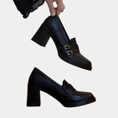 Paire de mocassins à talons hauts noirs, dont une chaussure est posée de profil et la deuxième tenue par les doigts d'une femme
